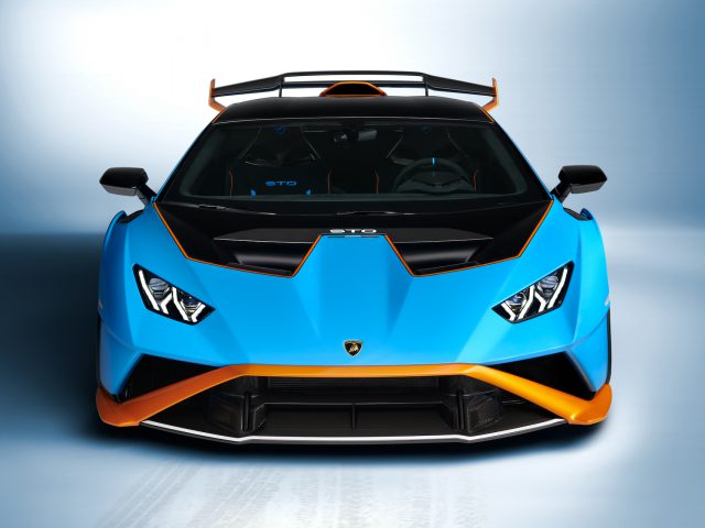 Vooraanzicht van een blauw-oranje Lamborghini Huracán STO met agressieve styling en LED-koplampen op een gradiëntblauwe achtergrond.