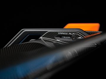 Close-up van de achterspoiler van een Lamborghini Huracán STO met koolstofvezeltextuur en een gecentreerde mediacamera, met een wazig oranje element op de achtergrond.