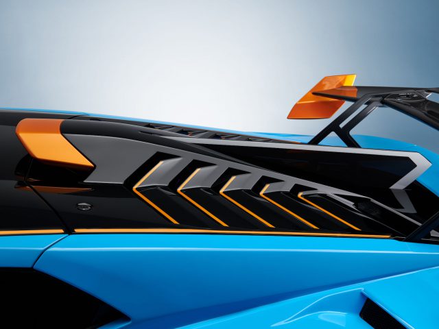 Close-up van een blauwe Lamborghini Huracán STO met oranje accenten, waarop de achterspoiler en ventilatieopeningen te zien zijn tegen een lichtblauwe achtergrond.