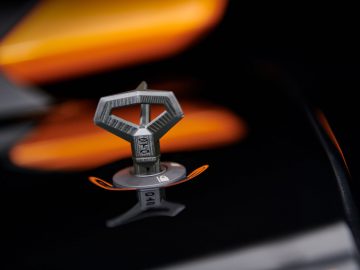 Close-up van een metalen autobadge in de vorm van een vlinderdas, gemonteerd op de grille van een Lamborghini Huracán STO met een wazige oranje motorkap op de achtergrond.