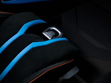 Close-up van een Lamborghini Huracán STO-autostoeltje met een veiligheidsgordelgesp in focus, gedetailleerd met blauwe en oranje accenten tegen een zwarte achtergrond.