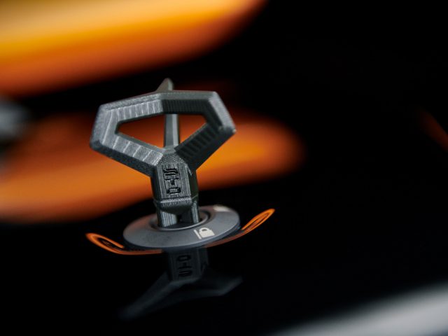 Close-up van een metalen badge op de motorkap van een auto, met een driehoekig logo met reflecterende oranje accenten, emblematisch voor de Lamborghini Huracán STO.