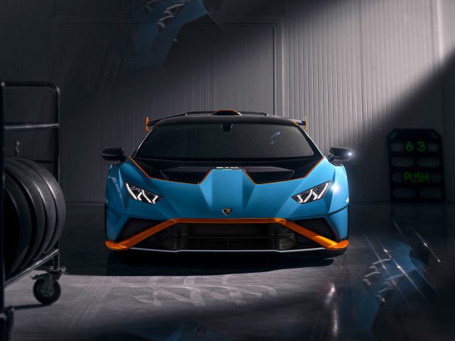 Een blauwe Lamborghini Huracán STO met oranje accenten geparkeerd in een slecht verlichte garage en scherpe schaduwen werpt op de glanzende vloer.