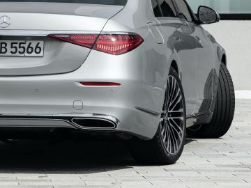 Achteraanzicht van een zilveren Mercedes-Benz S-Klasse met de achterlichten, de kofferbak en de kentekenplaat, geparkeerd op een betonnen ondergrond nabij een gebouw.