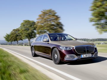 Een moderne Mercedes-Maybach S-Klasse in bordeauxrood rijdend op een zonnige weg met een helderblauwe lucht en groene bomen op de achtergrond.
