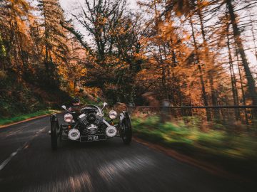 Een Morgan 3 Wheeler rijdt over een bosweg omringd door levendig herfstgebladerte.