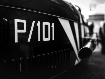 Close-up van het zijpaneel van een vintage Morgan 3 Wheeler, gemarkeerd met "p/101" met de nadruk op de nummering en de uitlaatpijp, onscherpe achtergrond.