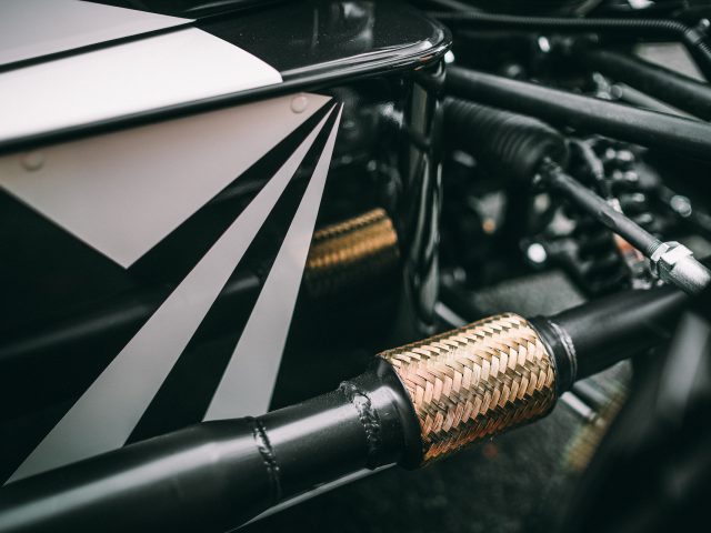 Close-up van een Morgan 3 Wheeler-uitlaatpijp met een hittebestendige hoes en gedetailleerde metalen onderdelen, met geometrische zwart-witte patronen op aangrenzende oppervlakken.