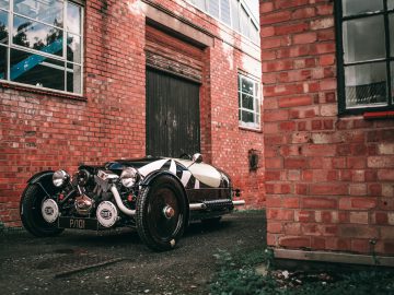 Een vintage Morgan 3 Wheeler geparkeerd naast een rood bakstenen gebouw met een grote houten deur.