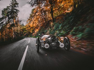 Een vintage Morgan 3 Wheeler die te hard rijdt op een bosweg omringd door herfstbomen.