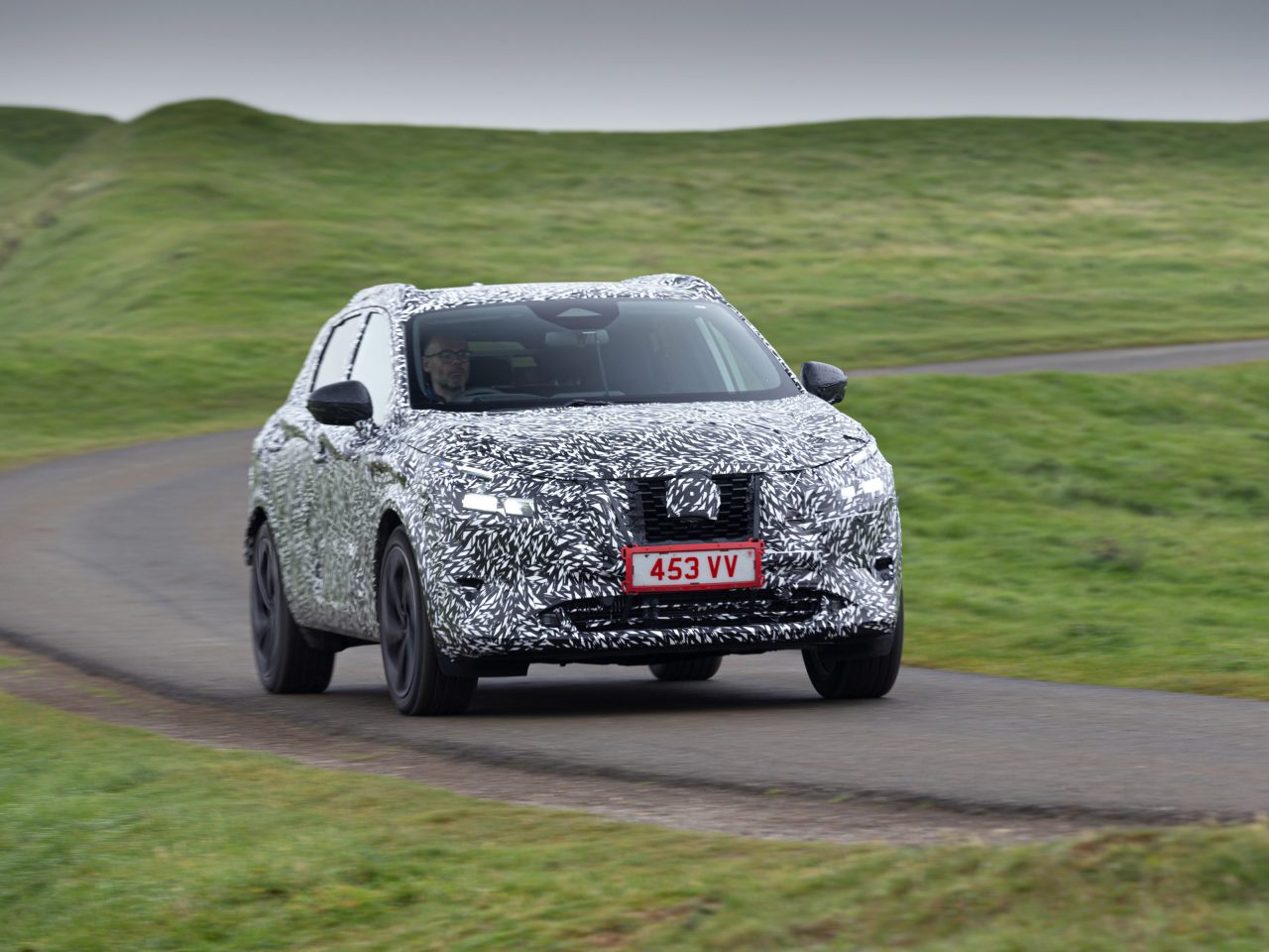 Een Nissan Qashqai met een gecamoufleerd ontwerp, rijdend op een bochtige weg met groene, met gras begroeide heuvels op de achtergrond.