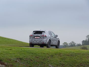 Een gecamoufleerd Nissan Qashqai-prototype rijdt op een bochtige weg in een met gras begroeid, heuvelachtig landschap onder een bewolkte hemel.
