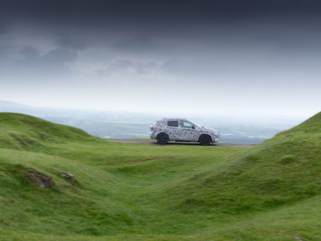 Een zilverkleurige Nissan Qashqai SUV geparkeerd op een met gras begroeide heuvel met donkere onweerswolken erboven, in een landelijk landschap.