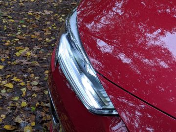 Close-up van de koplamp en motorkap linksvoor van een rode Opel Insignia, met gevallen herfstbladeren op de grond.