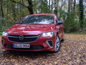 Rode Opel Insignia-auto geparkeerd op een met bladeren bedekte weg omringd door herfstbomen.