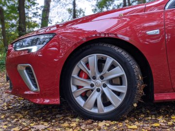 Close-up van de linkervoorzijde van een rode Opel Insignia, met het stuur en de koplamp, geparkeerd op een met bladeren bedekt pad in een bos.