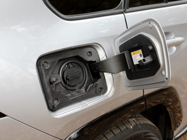 Close-up van een open tankdop op een zilveren Suzuki Across Plug-in Hybrid, met het mondstuk van de benzinepomp erin, met gedetailleerde labels en texturen.