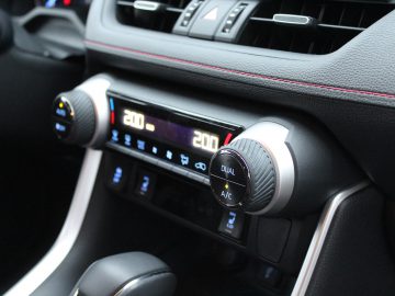 Close-up van het klimaatcontrolesysteem van een Suzuki Across Plug-in Hybrid met dubbele temperatuurinstellingen, digitale displays en zilveren knoppen.