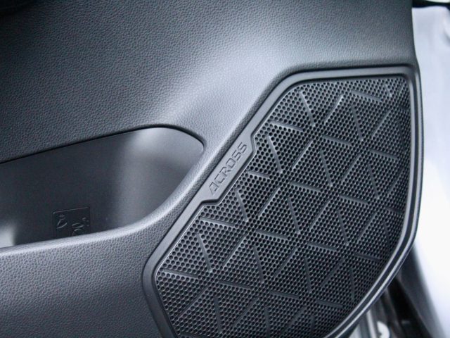 Close-up van de luidsprekergrille van een autodeur met geometrisch patroon en het opschrift 'Suzuki Across Plug-in Hybrid' op het zijpaneel.