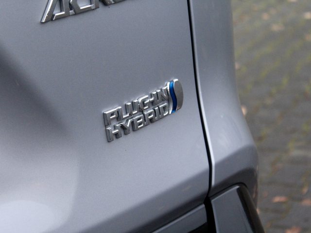 Close-up van een 'Suzuki Across Plug-in Hybrid'-badge op de achterkant van een grijze auto, die de hybridetechnologie van het voertuig aangeeft.