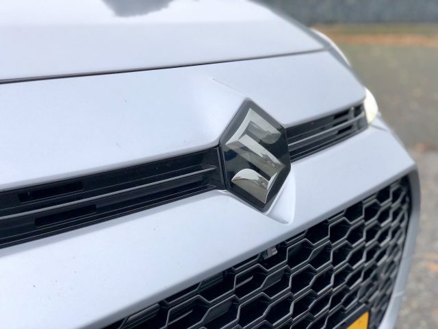 Close-up van de grille van een witte Suzuki Across Plug-in Hybrid met een opvallend logo, met de nadruk op scherpe details en een reflecterend oppervlak.