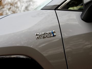 Close-up van een 'Suzuki Across Plug-in Hybrid'-badge op het spatbord van een witte auto, met de nadruk op de details en textuur van het metalen embleem.