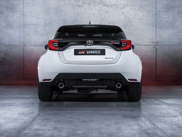 Achteraanzicht van een Toyota GR Yaris, geparkeerd voor een muur met grijze textuur, met de badges en dubbele uitlaatpijpen.