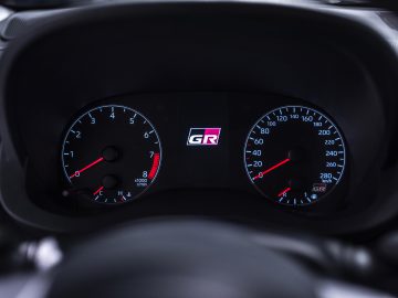 Autodashboard met een toerenteller en een snelheidsmeter met een "Toyota GR Yaris"-logo, verlicht tegen een donkere achtergrond.