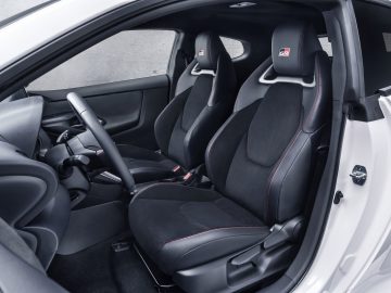 Binnenaanzicht van een witte Toyota GR Yaris-sportwagen met twee voorstoelen met zwarte bekleding gemarkeerd met het GR-logo, het stuur, de versnellingspook en de bedieningselementen op het dashboard.