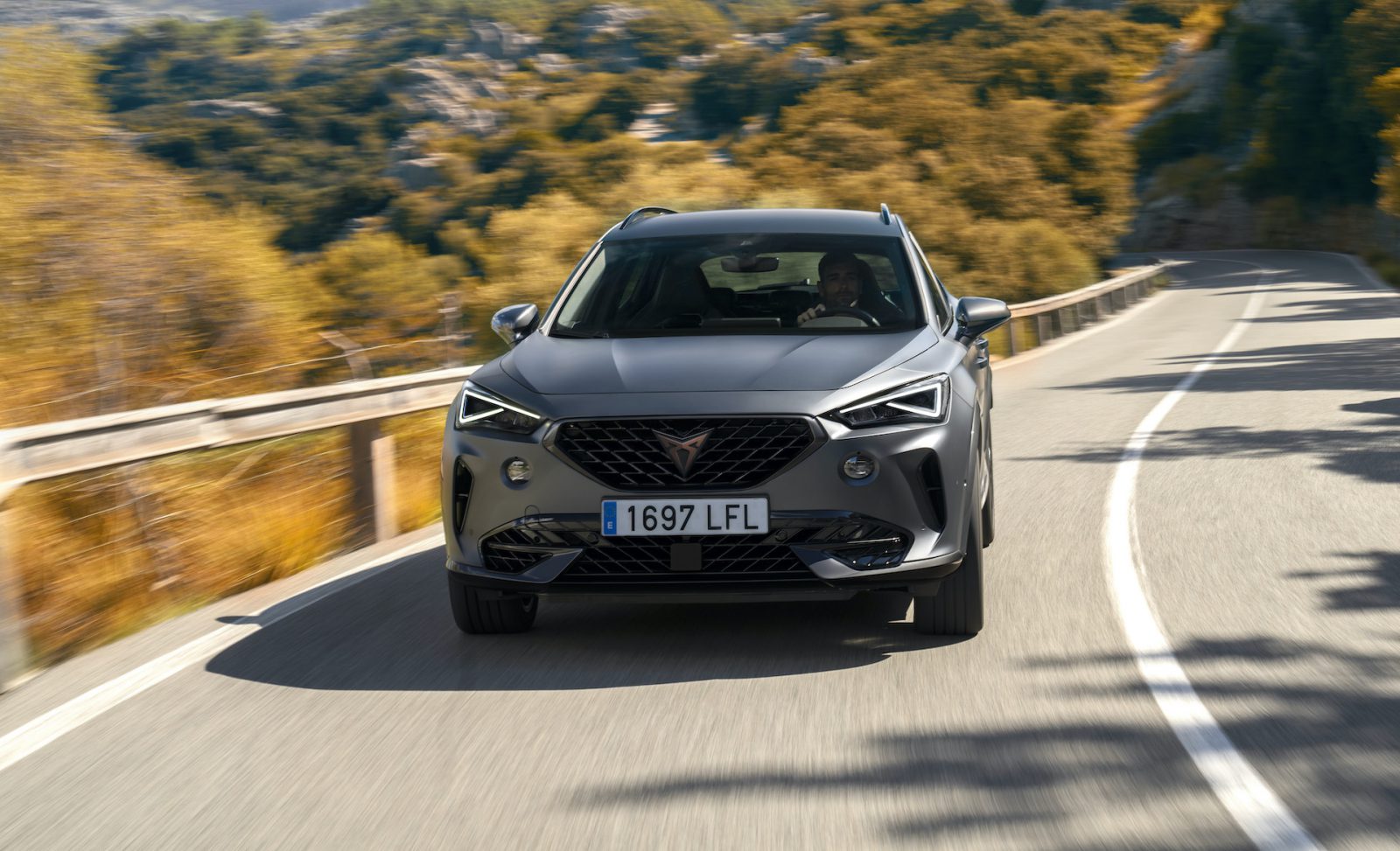 Een grijze Cupra Formentor SUV die over een zonovergoten bergweg rijdt, waarbij de kenmerkende grille en strakke koplampen prominent tot hun recht komen.