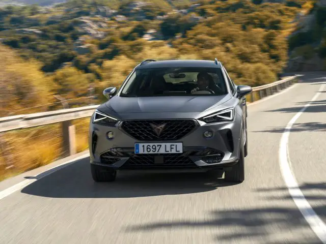 Een grijze Cupra Formentor SUV die over een zonovergoten bergweg rijdt, waarbij de kenmerkende grille en strakke koplampen prominent tot hun recht komen.