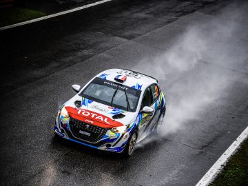 Een Peugeot 208 Rally 4-raceauto met een kleurrijke kleurstelling snelt op een natte baan, waardoor er een waterstraal achter ontstaat.