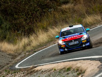 Een Peugeot 208 Rally 4-auto, voorzien van 'totale' sponsorstickers, snelt een bocht door op een onverharde bergweg en doet stof opwaaien.