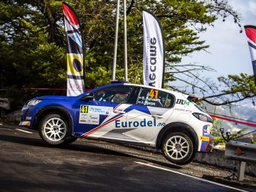 Een Peugeot 208 Rally 4-auto gemarkeerd met nummer 41 sponsors, waaronder "eurodel.no", die over een gebarricadeerde weg racet met fladderende promotievlaggen op de achtergrond.