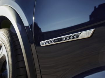 Close-up van het ventilatierooster aan de zijkant van een DS 7 CROSSBACK met het woord "louvre" en een geometrisch patroon, net boven het voorwiel op een donkerblauwe carrosserie.