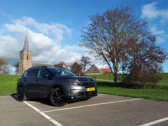 Een zwarte C5 Aircross Hybrid geparkeerd op een parkeerplaats met een oude kerk en een gedeeltelijk bewolkte lucht op de achtergrond.
