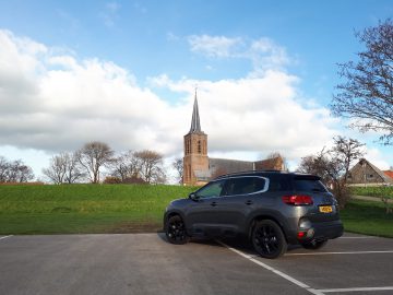 Een zwarte C5 Aircross Hybrid geparkeerd voor een met gras begroeide dijk met een historische kerk met een hoge torenspits op de achtergrond onder een gedeeltelijk bewolkte hemel.
