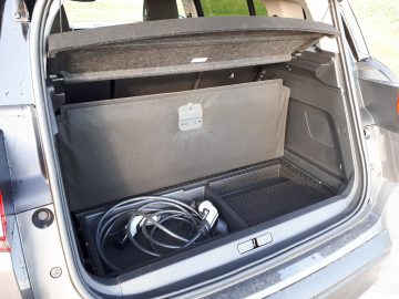 Open kofferbak van een C5 Aircross Hybrid met een opbergvak met kabel en een beschermmat, gezien bij daglicht.