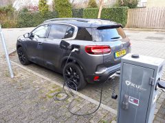Een grijze C5 Aircross Hybrid is overdag aangesloten op een openbaar laadstation op een parkeerplaats.
