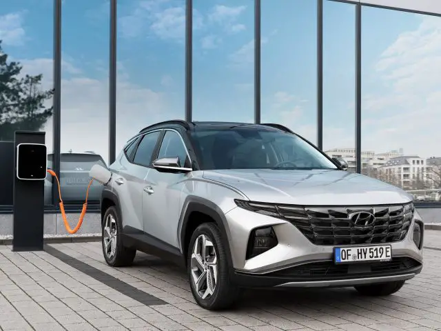 Zilverkleurige Hyundai Tucson Plug-in Hybrid SUV geparkeerd voor een glazen gebouw, aangesloten op een elektrisch laadstation.