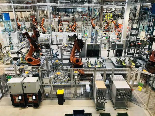 Geautomatiseerde fabrieksvloer van INEOS Automotive met meerdere robotarmen die onderdelen assembleren, omgeven door veiligheidshekken en machines.