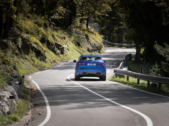 Een blauwe Jaguar F-Pace SVR rijdt op een bochtige bergweg omzoomd met bomen en vangrails onder een zonnige hemel.