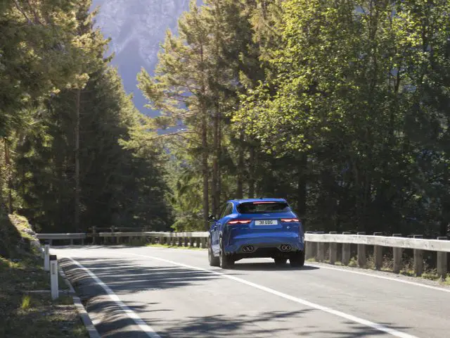 Een blauwe Jaguar F-Pace SVR die over een bochtige bergweg rijdt, omringd door dichte groene bomen en een helderblauwe lucht.