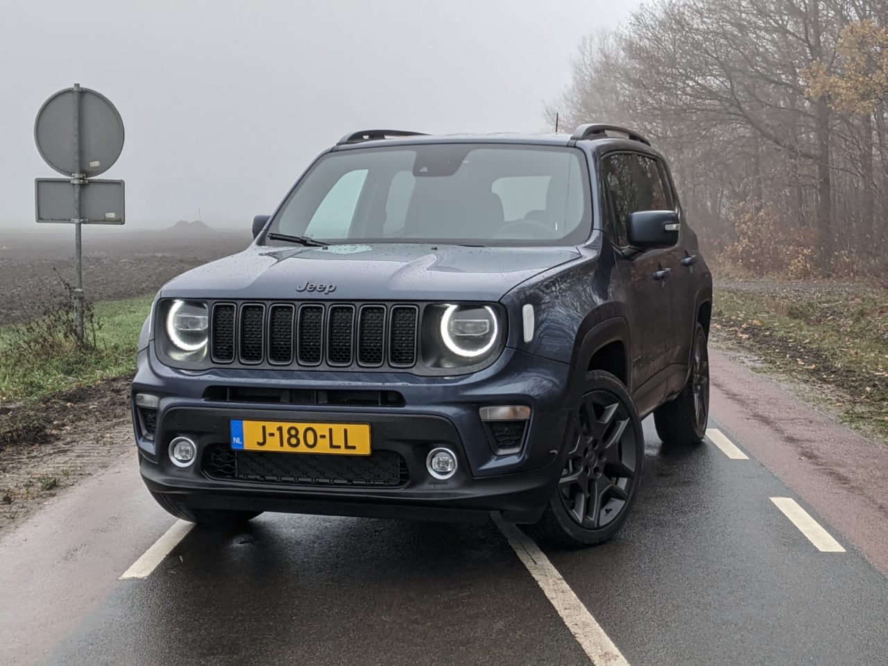 Een blauwe Jeep Renegade 4xe geparkeerd op een mistige weg met koplampen aan, omgeven door een mistig landelijk landschap.