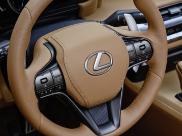 Close-up van een Lexus LC 500 Convertible-stuurwiel met bruin leer en ingebouwde bedieningselementen, tegen een dashboard met digitale displays.