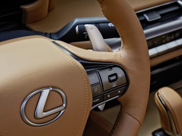 Close-up van een Lexus LC 500 Convertible-stuurwiel met bedieningsknoppen, met een deel van het dashboard en het beige lederen interieur.