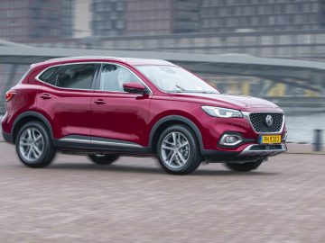 MG EHS Plug-in Hybrid rijdt op een bewolkte dag door een stadsstraat in de buurt van gebouwen aan het water.