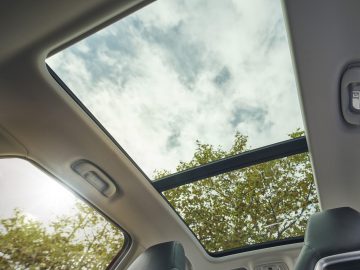 Uitzicht op een lucht met wolken door een open zonnedak van een MG EHS Plug-in Hybrid-auto, omlijst door het interieur van het voertuig, inclusief zonnekleppen en dakgrepen.