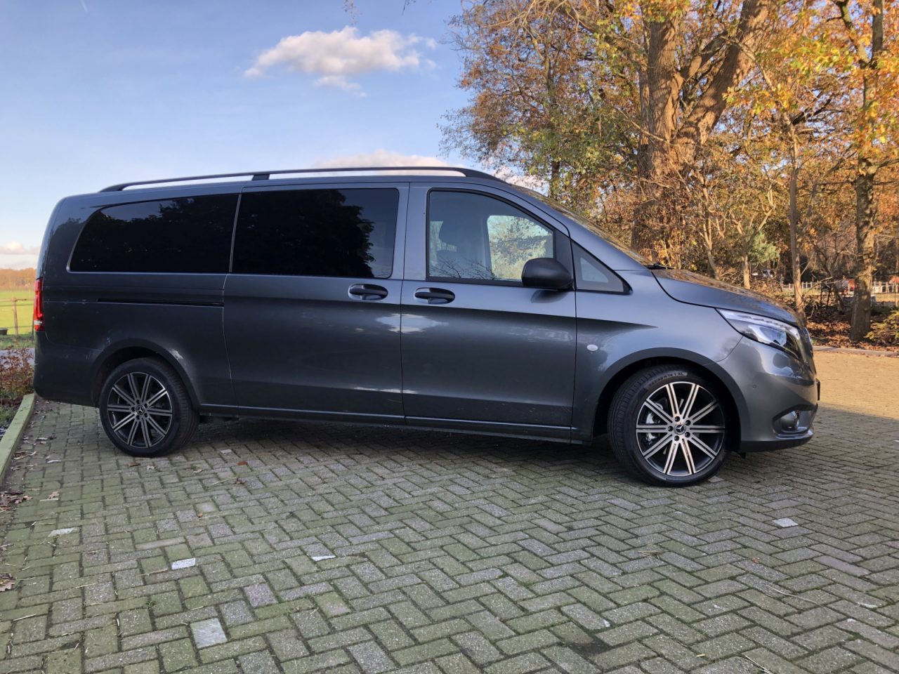 Een grijze Mercedes-Benz Vito geparkeerd op een stenen oprit met bomen en een grasveld op de achtergrond op een zonnige dag.