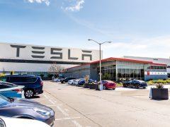 Buitenaanzicht van een Tesla-faciliteit met grote bewegwijzering, een parkeerplaats vol auto's en een helderblauwe lucht.